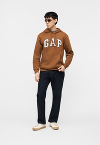 Νέος άνδρας που φοράει καφέ hoodie GAP, σκούρο τζιν, λευκά αθλητικά παπούτσια και γυαλιά ηλίου, στέκεται μπροστά σε ένα απλό φωτεινό φόντο.