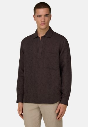 Mężczyzna w ciemnobrązowej koszuli z długim rękawem i kieszenią na piersi oraz beżowych spodniach, stojący na tle jednolitego, jasnego tła.