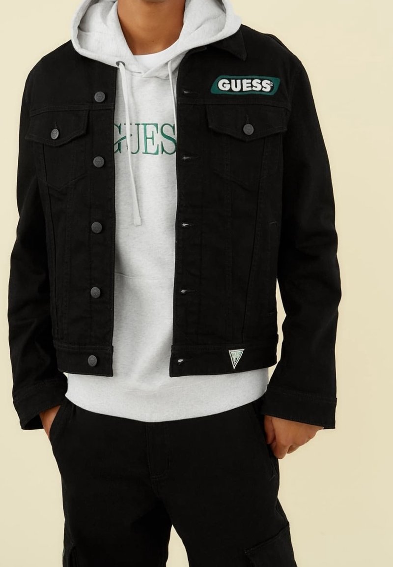 Svart denimjacka med knappfront, två bröstfickor och logopatcher. Bärs över en grå hoodie med grön text. Avslappnad outfit.