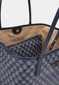 Granatowa torba na ramię z geometrycznym wzorem, z zamykaną kieszonką w środku. Podszewka z beżowego materiału, wytłoczona nazwą marki.