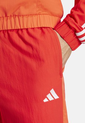 Pantalones deportivos rojos con una cintura elástica, bolsillos laterales y el logo de Adidas en blanco. Presentan una textura ligera y suave, con acentos en naranja contrastante.