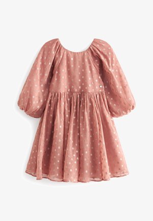 Junges Mädchen in einem pinkfarbenen Kleid mit goldenen Punkten und Puffärmeln, trägt metallisch goldene Sandalen, steht vor einem schlichten Hintergrund.
