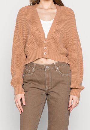 Femme portant un cardigan court en grosse maille beige avec trois boutons sur un top blanc, assorti à un pantalon marron taille haute.