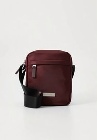 Sac bandoulière en tissu bordeaux de forme rectangulaire, avec une sangle noire réglable, une fermeture éclair, et un logo métallique en accent sur le devant.