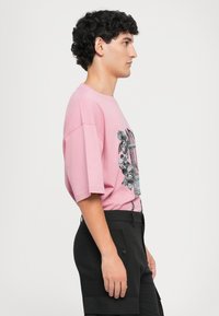 Ροζ oversized μπλουζάκι με μαύρο γρ�αφικό σχέδιο που απεικονίζει λουλούδια και ένα κλουβί. Το μοντέλο φοράει μαύρες βερμούδες, άποψη από το πλάι.