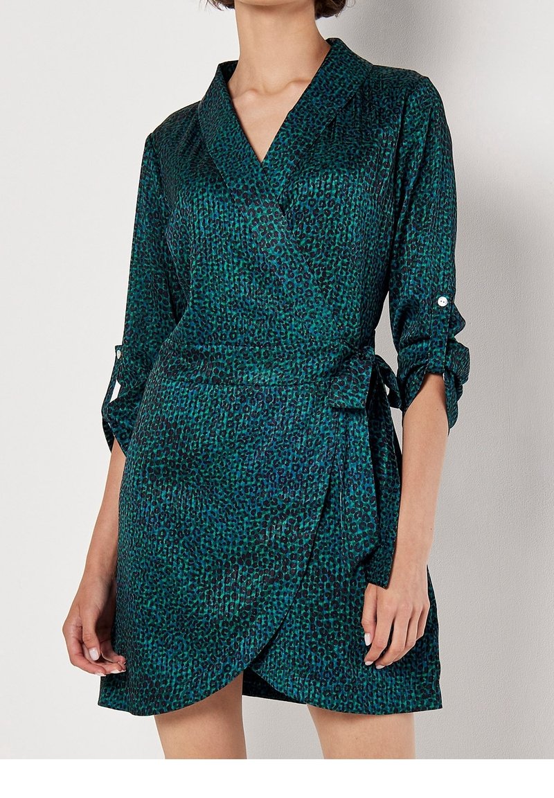 Femme portant une robe portefeuille imprimée animalier turquoise et noire, manches trois-quarts retroussées et ceinture nouée sur le côté, debout devant un fond uni.