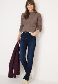 Pull marron en maille torsadée, jean ajusté bleu foncé, tenant une veste texturée bordeaux. Chemise blanche en dessous, baskets de couleur claire.