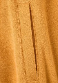 Prim-plan al unei țesături moi, texturate, din bumbac tip prosop, de culoare muștar-galben, cu un buzunar dreptunghiular vertical cusut pe ea.