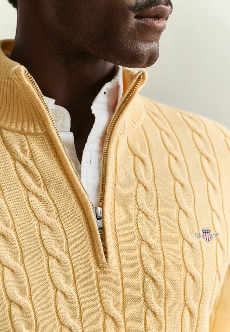 Man wearing a yellow cable-knit zip-up sweater over a white button-up shirt, showing his lower face and neck.