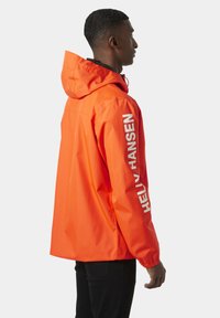 Oranje waterdichte jas met capuchon, voorzien van witte tekstbranding op de rechter mouw en elastische manchetten voor een comfortabele pasvorm.