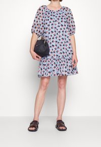 Kobieta ubrana w jasnoniebieską sukienkę w kwiaty z bufkami na rękawach i falbanką na dole, trzymająca czarną torebkę i nosząca ciemnobrązowe sandały.