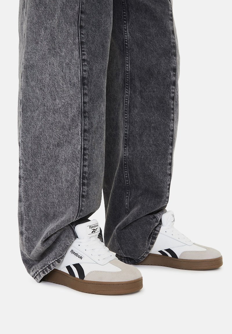 Białe i szare sneakersy Reebok z czarnymi akcentami, w połączeniu z luźnymi, ciemnoszarymi dżinsami. Sneakersy mają gumową podeszwę i są sznurowane.