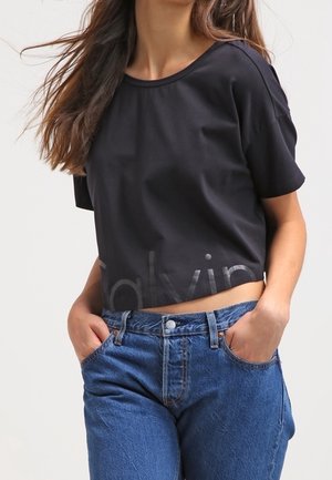 Woman wearing a black cropped t-shirt with text and high-waisted blue jeans, standing with hands in pockets against a plain background.