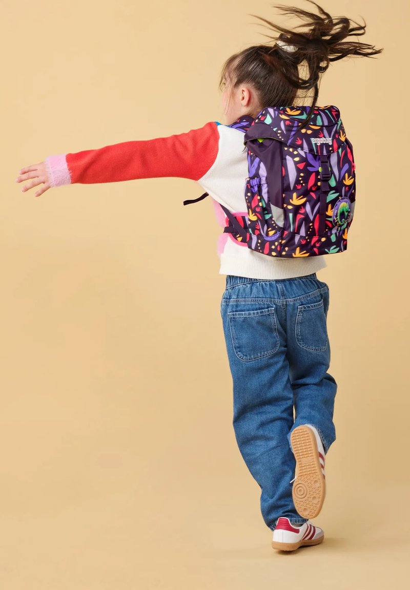 Kind met lang haar in beweging, gekleed in een kleurrijke bloemenrugzak, een rood-wit sweater, blauwe jeans en witte sneakers op een beige achtergrond.