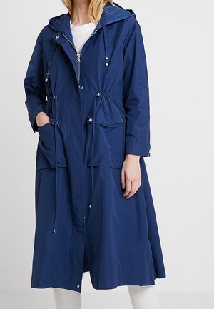 Femme portant un long imperméable à capuche bleu marine avec fermeture éclair à l'avant, taille à cordon de serrage, poches à boutons-pression, et pantalon blanc.
