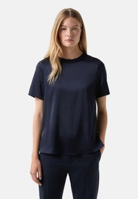 Marineblaues Kurzarm-T-Shirt aus glattem Stoff mit rundem Ausschnitt, in lockerer Passform und leichtem Glanz. Kombiniert mit dunklen Hosen.