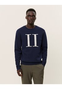 Sudadera azul marino con un gráfico de felpa blanca "II" en la parte delantera, cuello redondo y puños acanalados. Combinada con pantalones de color claro.