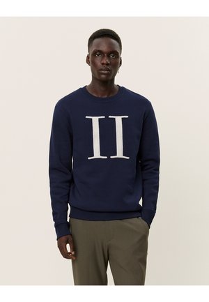 Sudadera azul marino con un gráfico de felpa blanca "II" en la parte delantera, cuello redondo y puños acanalados. Combinada con pantalones de color claro.