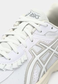 Gros plan d'une basket blanche Asics en tissu mesh, avec des lacets blancs et un motif géométrique gris sur le côté.