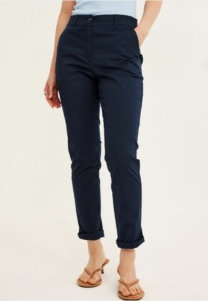 Nainen pukeutunut laivastonsinisiin kapeisiin, käännettyihin housuihin ja ruskeisiin korollisiin sandaaleihin, seisoo yhdellä kädellä taskussa yksivärisen taustan edessä.