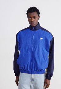 Zils un tumši zils ņiprs vēja necaurlaidīgs apmetnis ar elastīgām piedurknēm un jostu, ar balto Nike logotipu uz krūtīm. Viegls, gluds audums.