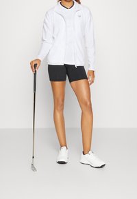 Bílá zipová bunda, černé přiléhavé šortky, držící golfovou hůl. Bílé sportovní boty s černými akcenty. Hladká látka, sportovní střih.