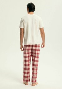 Biała koszulka z krótkim rękawem w połączeniu z czerwono-kremowymi, w kratę spodnie do piżamy. Miękka tkanina o luźnym kroju. Widoczne bose stopy.