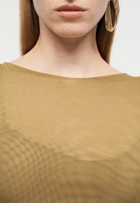 Κοντινό πλάνο του κατώτερου μέρους του προσώπου και του λαιμού ενός ατόμου που φοράει ένα διαφανές τοπ μουσταρδί χρώματος με ένα μεγάλο χρυσό κρίκο σκουλαρίκι ορατό.