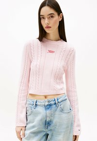 Suéter corto de punto en rosa claro con patrones de trenzas y puños acanalados, con un logo rojo de "Tommy Jeans" en el pecho. Combinado con jeans azules.