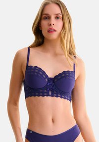 Soutien-gorge violet en tissu ribbé avec des détails en dentelle. Présente des bonnets structurés, des bords festonnés et un petit détail en forme de trou de serrure à l'avant.