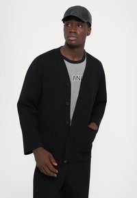 Crni kardigan s prednjim gumbima, V-izrezom i bočnim džepovima, u kombinaciji sa sivom majicom. Muškarac nosi crnu kapu, neutralna pozadina.