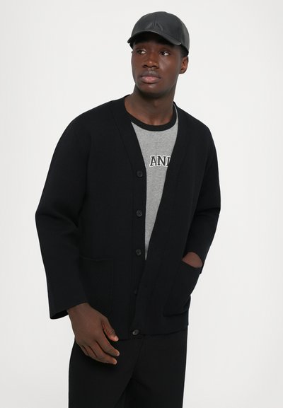 Musthave mustärmine kardigan nööbiga ees, V-kaelusega ja küljetaskutega, koos halli T-särgiga. Mees kannab musta mütsi, neutraalne taust.