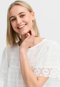 Top bianco realizzato in tessuto testurizzato con un motivo a griglia; presenta maniche corte a sbuffo e delicati pizzi decorativi ai polsini.