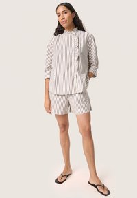 Stribet kortærmet skjorte med volangdetalje og knaplukning, parret med matchende stribede shorts. Farverne er creme og brun.