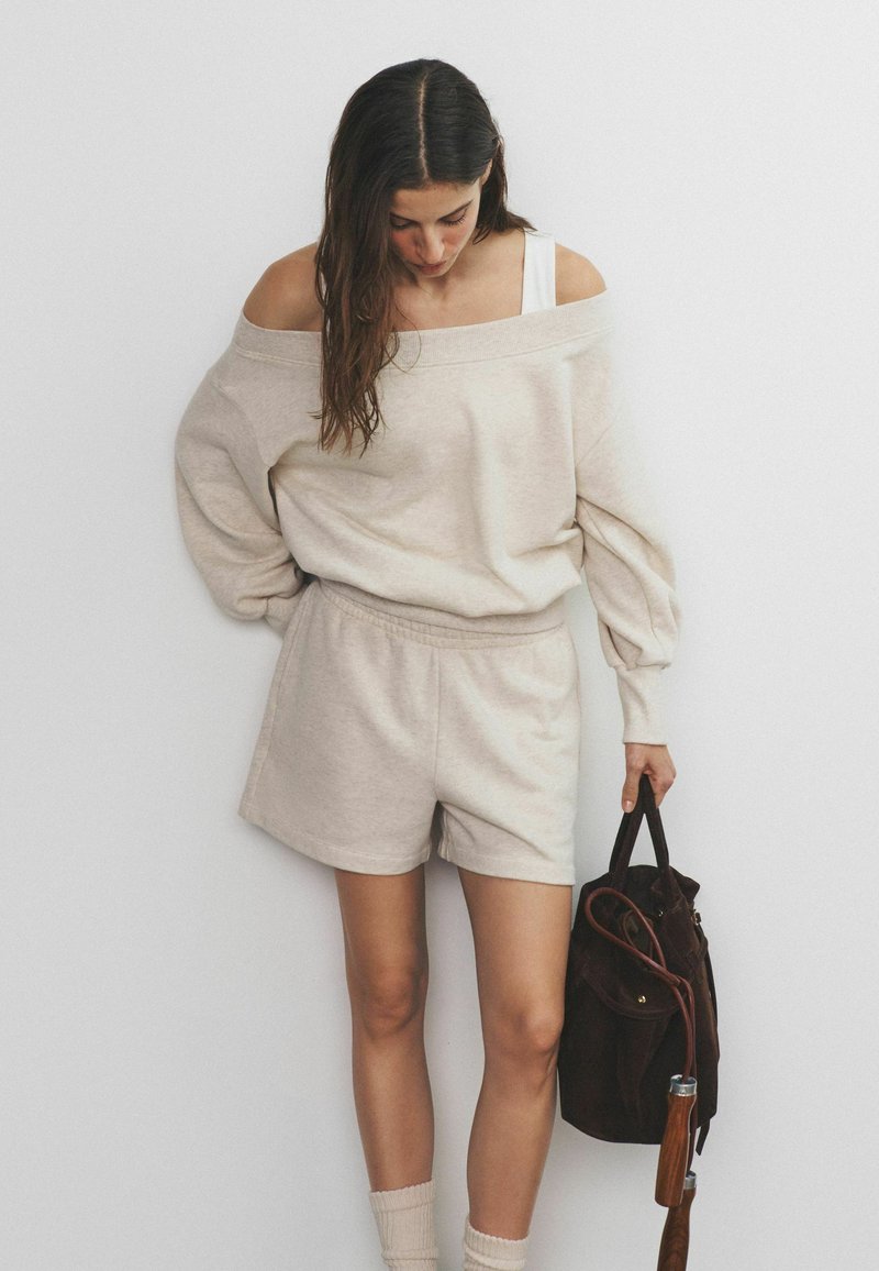 Vrouw die omlaag kijkt, draagt een beige off-shoulder trui en korte broek, en houdt een donkerbruine suède tas met houten handvatriemen vast.