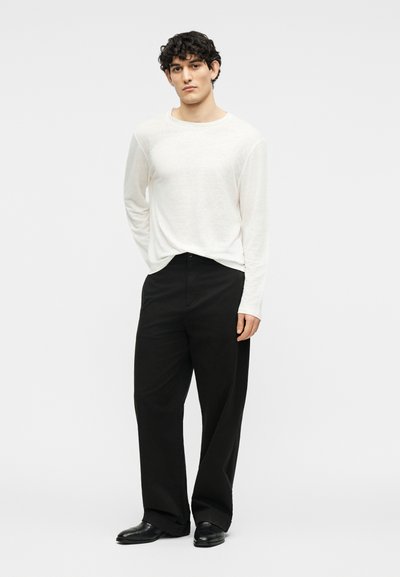 Άνδρας που στέκεται φορώντας λευκό πουκάμισο με μακριά μανίκια, φαρδιά μαύρα παντελόνια και μαύρα επίσημα παπούτσια, μπροστά σε απλό ανοιχτόχρωμο φόντο.