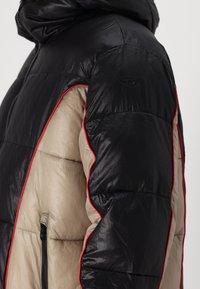 Casaco puffer com exterior preto e interior bege, realçado com acabamentos em vermelho. Inclui um bolso com fecho éclair e capuz.
