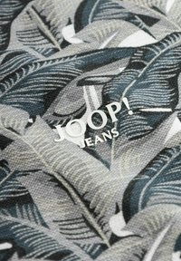 Tissu à motifs présentant des feuilles grises et noires sur un fond clair ; logo "JOOP! JEANS" en blanc, positionné au centre.