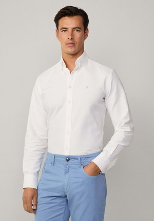 Camicia bianca con colletto, caratterizzata da un logo azzurro sul petto, abbinata a chinos azzurri. Tessuto morbido, design aderente.