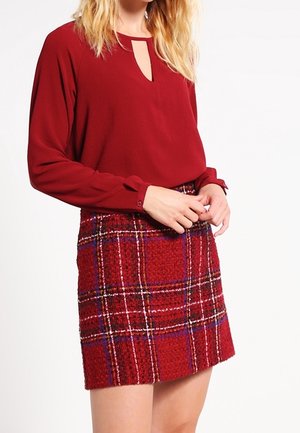 Rode blouse met lange mouwen en een keyhole-halslijn, gecombineerd met een geruite, gestructureerde rode rok met blauwe en witte accenten.