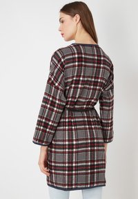 Bata de cuadro en gris, rojo y negro. Hecha de una tela suave con mangas largas, cintura con cinturón y un ribete en contraste de color marino a lo largo del escote.