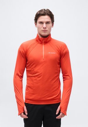 Noor mees, kes kannab erkoranž Columbia pika varrukaga sportlikku särk, millel on pöidlaaugud, seisab ühevärvilise heleda tausta ees.