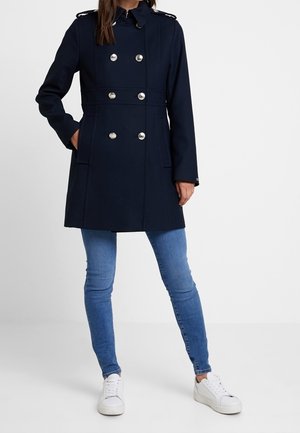 Kvinde iført en marineblå dobbeltradet frakke, lyseblå skinny jeans og hvide sneakers, stående med den ene hånd i lommen.