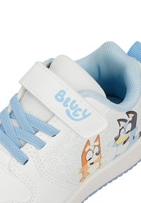 Baskets blanches pour enfants avec des lacets bleu clair, une bride velcro étiquetée « Bluey » et des personnages de dessins animés sur le côté.