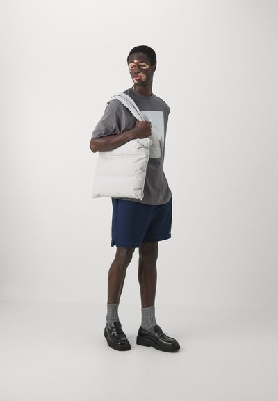 Gevoerde witte schoudertas vastgehouden door een persoon in een grijs shirt en marineblauwe shorts, die een minimalistisch ontwerp en een gladde textuur toont.