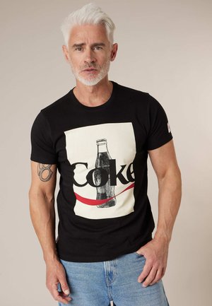 Homme aux cheveux blancs et à la barbe portant un t-shirt noir avec un motif vintage de bouteille de Coca-Cola et un jean bleu clair, debout devant un fond uni.