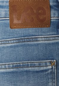 Etiqueta de cuero marrón con la marca Lee cosida en una tela vaquera azul claro, con costuras doradas y un diseño clásico de cinco bolsillos.