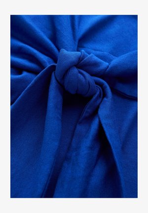 Sinine puuvillane kangas sõlmega keskel, sile tekstuur ja kergelt venitatud alad, mis loovad sõlme ümber kogutud efekti.