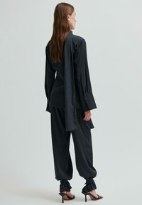 Czarna satynowa bluzka z długim tylnym szalem, zestawiona z luźnymi czarnymi spodniami. Ubranie charakteryzuje się wyrazistymi mankietami i związanymi detalami w okolicy kostek.