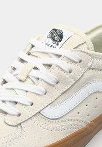 Beige semsket sneaker med hvit skinnstripe på siden, hvite snørebånd og gummisåle. Har merket etikett på tungen og perforerte detaljer.